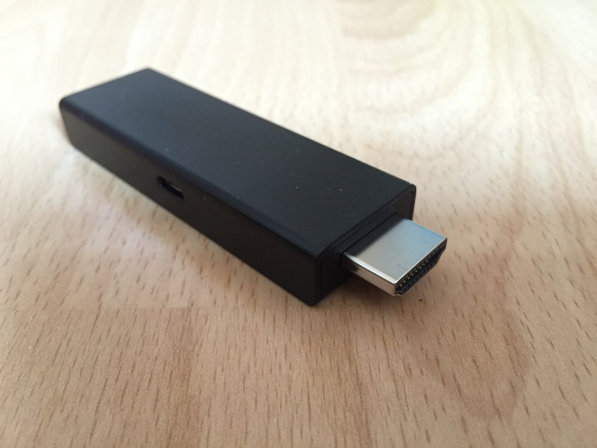 Amazon Fire TV Stick inkopplad i en TV:s HDMI-port med fjärrkontrollen framför en startskärm med strömningsappar